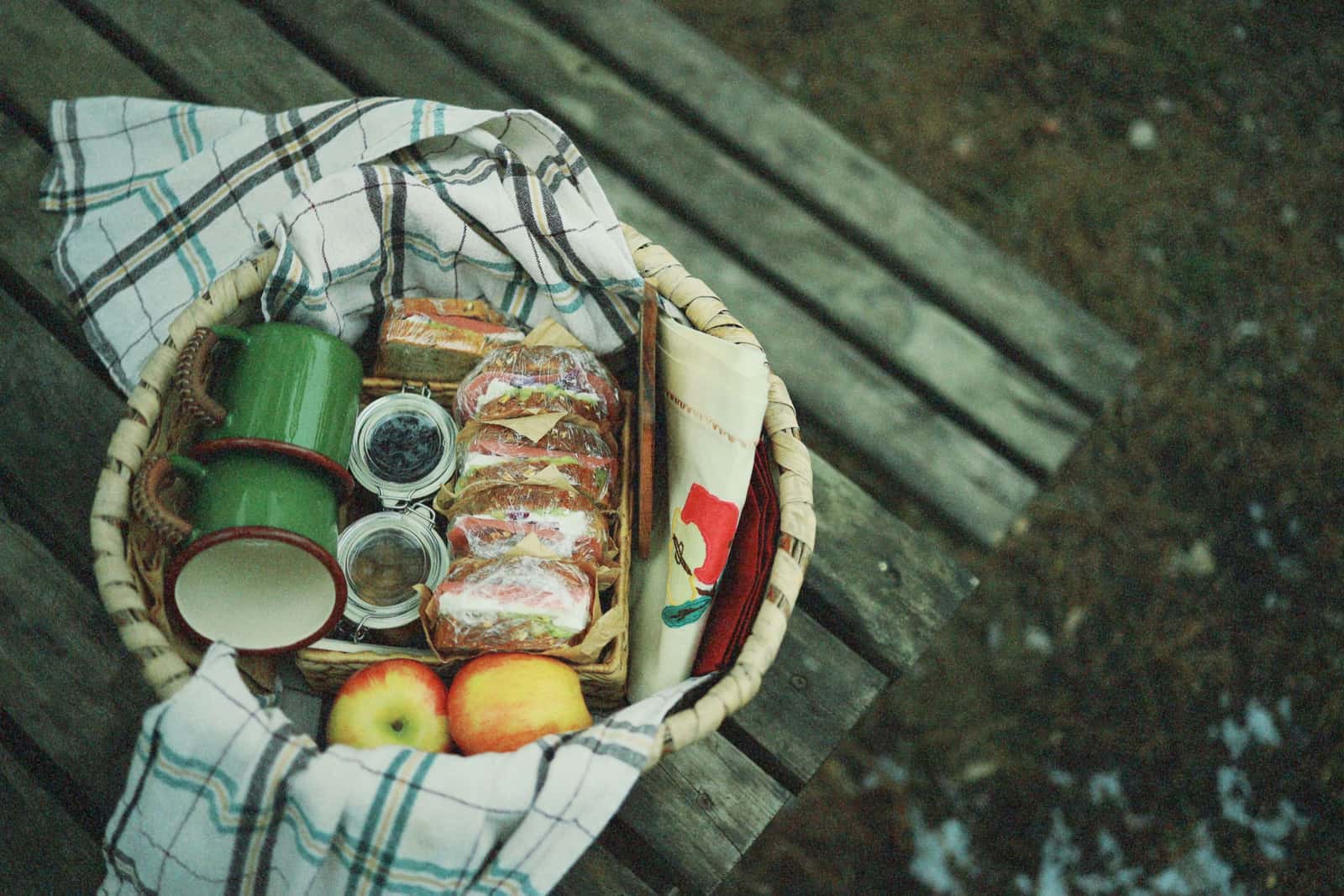 A picnic basket with sandwiches and apples on a wooden bench