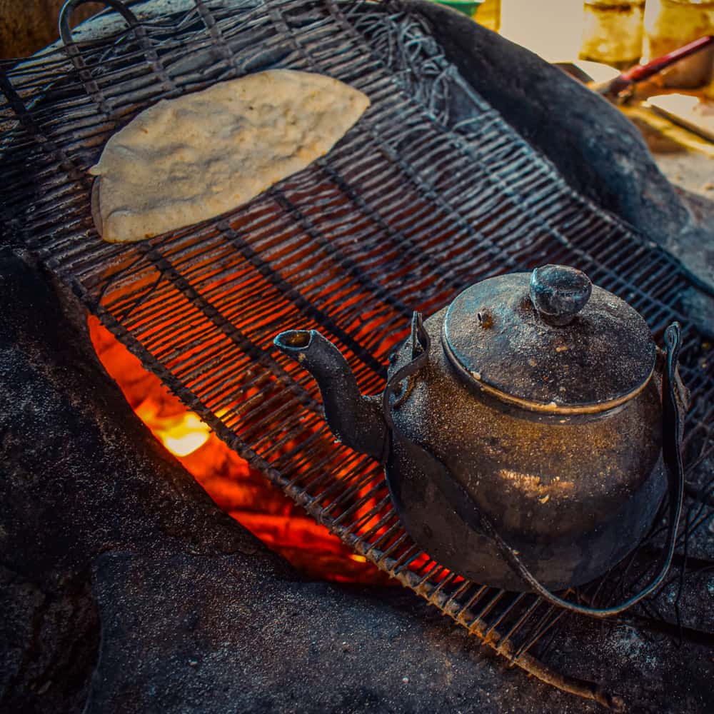 Illustrative stock photo: a kettle and flatbread cooking over an open campfire grill