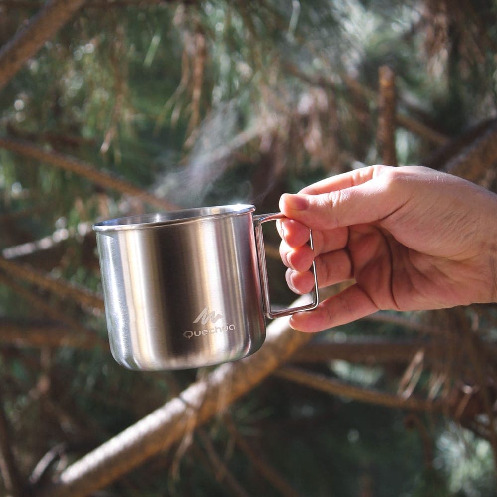Illustrative stock photo: a hand holding a steaming steel mug outdoors in a forest
